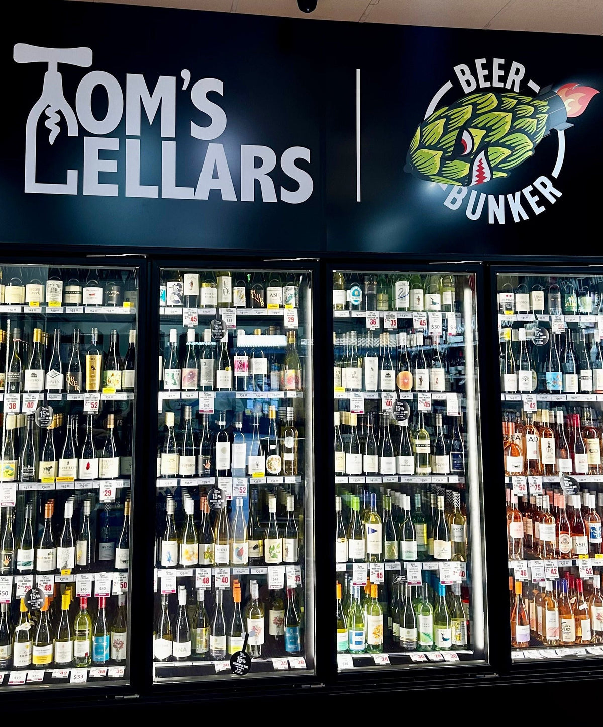 A retail display of Tom's Lellars wine and beer refrigerated glass-door cases under a branded black sign. The shelves are stocked with numerous wine bottles and beer bottles, and the floor is wood-look laminate.