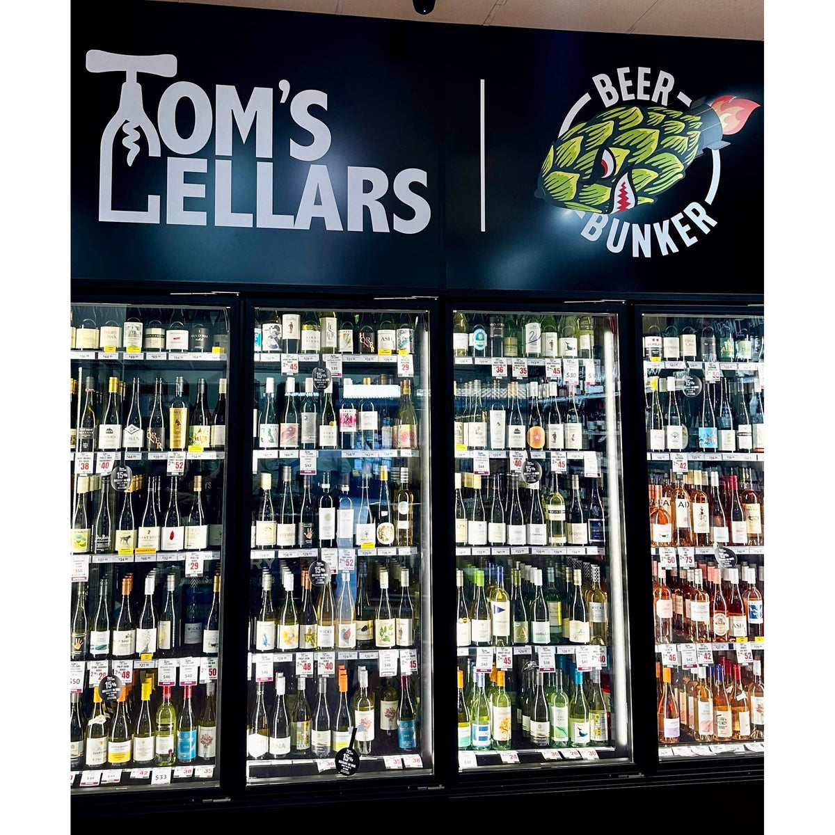 A retail display of Tom's Lellars wine and beer refrigerated glass-door cases under a branded black sign. The shelves are stocked with numerous wine bottles and beer bottles, and the floor is wood-look laminate.